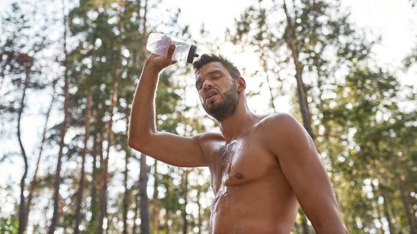 Exhausted young caucasian sportsman pouring water on himself from bottle in blurred forest. Handsome bearded man with naked torso and closed eyes. Concept of modern healthy lifestyle. Warm summer day - Fotografie, Obrázek