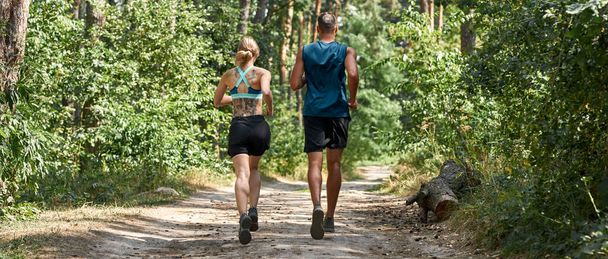 Back view of young sports couple jogging on path in green forest. Pretty blonde slim woman with tattoos and handsome man wearing sportswear. Concept of modern healthy lifestyle. Sunny summer day - Fotó, kép