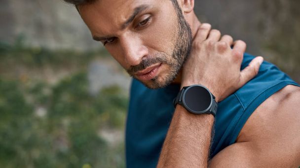 Cropped view of young caucasian sportsman thinking about something outdoors. Handsome bearded athletic man wearing sportswear and smartwatch. Concept of modern healthy lifestyle. Warm summer day - Foto, Imagen
