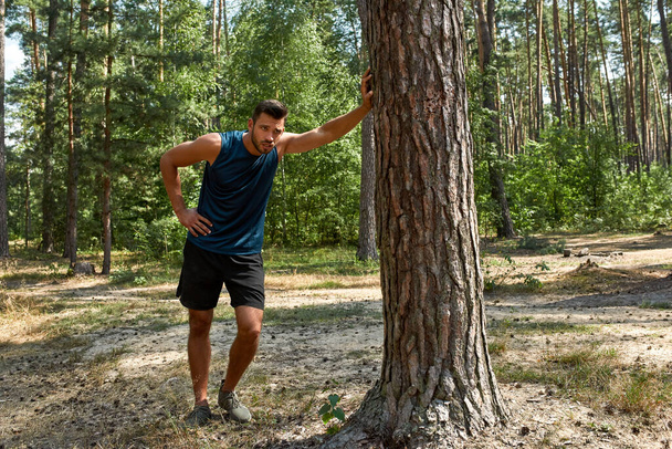 Tired young caucasian sportsman with hand on hip touching tree and resting after running in green forest. Handsome bearded man wearing sportswear. Concept of modern healthy lifestyle. Sunny summer day - Φωτογραφία, εικόνα