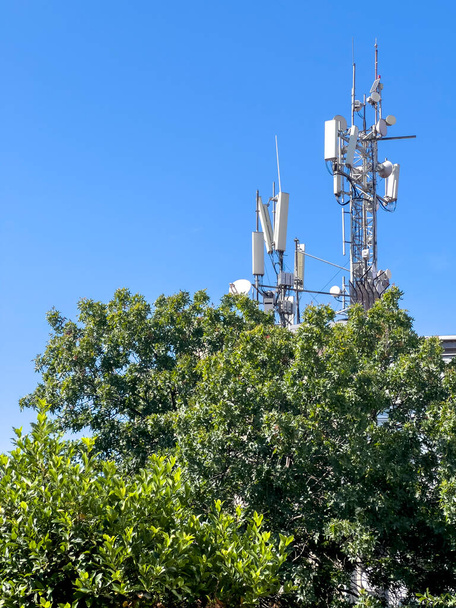GSM Antenna Torony, másold a helyet. Bázisadó állomás. Vezeték nélküli telekommunikációs technológia berendezés 5G, 4G, 3G. Mobiltelefon, TV antennák a tetőn zöld fa. Sunny ég háttér. - Fotó, kép