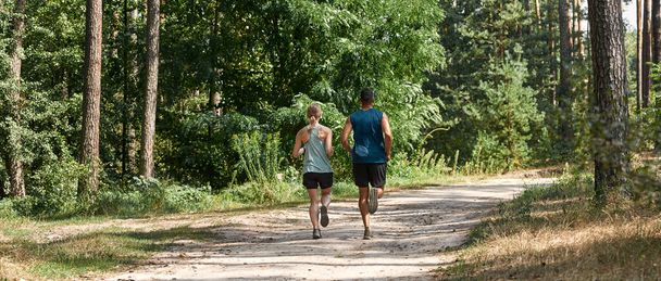 Back view of young sports couple running on path in green forest. Beautiful blonde slim woman with tattoos and handsome man wearing sportswear. Concept of modern healthy lifestyle. Sunny summer day - Foto, imagen