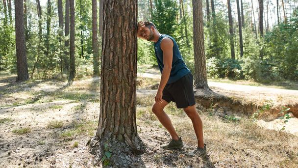 Tired young european sportsman touching tree and resting after running in green forest. Handsome bearded dark-haired man wearing sportswear. Concept of modern healthy lifestyle. Sunny summer day - Foto, imagen
