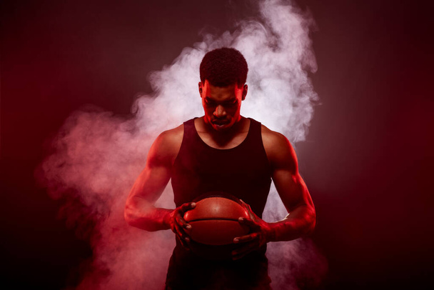 Basketball player side lit with red color holding a ball against smoke background. Serious concentrated african american man. - Фото, зображення