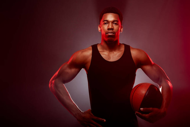 Basketball player side lit with red color holding a ball against hazy background. Serious concentrated african american man. - 写真・画像