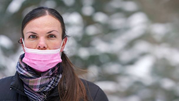 Young woman in winter jacket with pink disposable single use face virus mask improperly - nose not covered, space for text (blurred background snow covered trees) right side - Фото, зображення