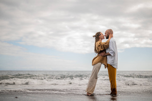 Młoda kochająca się para stojąca i przytulająca się na plaży. Obraz pokazuje intymność związku małżeńskiego. Wygląda na to, że ta para zaraz się pocałuje. Wysokiej jakości zdjęcie. Chmurna pogoda. - Zdjęcie, obraz