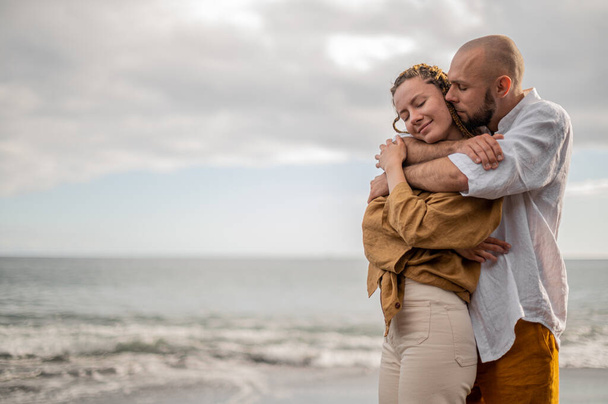 Young loving couple standing and hugging on a beach. Image is showing intimacy of the couple relationship. It seems that couple is about to kiss. High quality photo. Cloudy weather. - Photo, Image