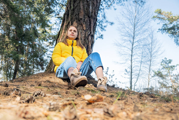 Portret uśmiechniętej młodej dziewczyny siedzącej w lesie pod drzewem. zdrowie psychiczne. Kobieta nosi jasnożółtą kurtkę. Czas dla siebie, restart, medytacja. - Zdjęcie, obraz