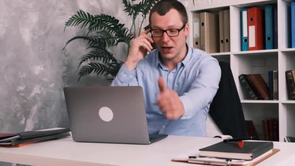 Empresário bem sucedido, gerente com óculos está falando em um telefone celular enquanto sentado na mesa de trabalho e laptop no fundo do rack com documentos e pastas no escritório ou em casa. - Filmagem, Vídeo