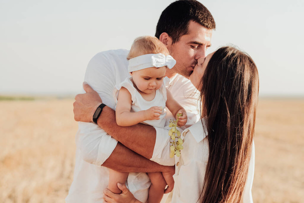 Nuori valkoihoinen perhe, jolla on lapsi, joka halailee ulkona pellolla, äiti ja isä suutelevat, kun heidän tyttärensä syö rypäleen. - Valokuva, kuva