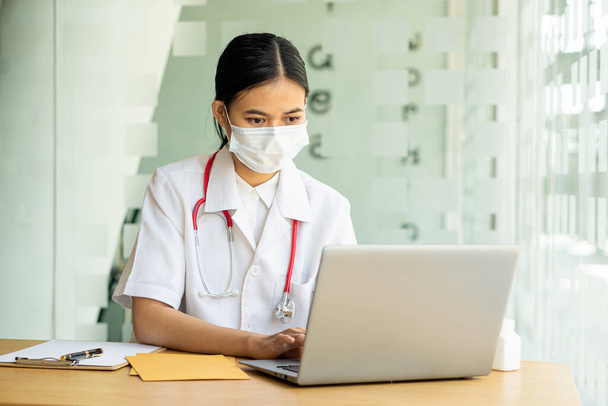 Asyalı bir kadın doktor hastane masasında çalışıyor, hastalara uygun online hizmetler öneriyor ve sağlık hizmeti tavsiyeleri içeren bir ilaç şişesi tutuyor. hastalık önleme kavramı - Fotoğraf, Görsel