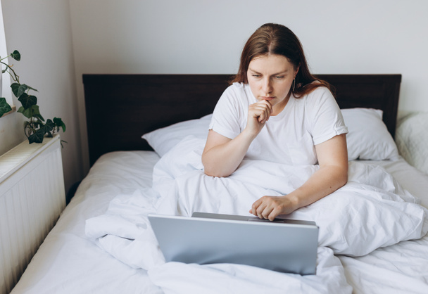 Stresszes, fiatal, vörös hajú nő laptoppal az ágyon egy lusta napon. Online oktatás, munka vagy vásárlás. Technológia női koncepció alternatív iroda, szabadúszó - Fotó, kép