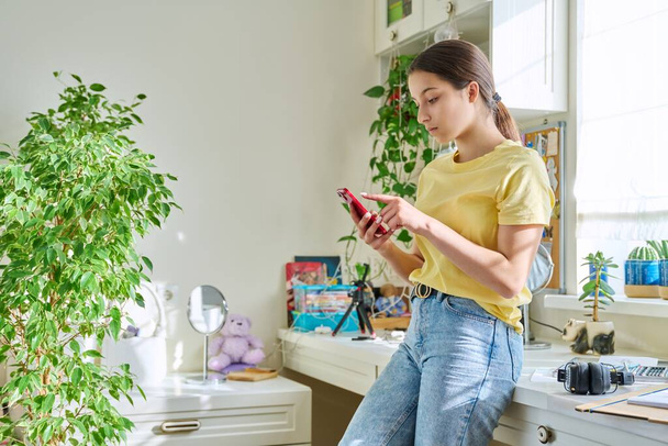 Happy teenage female using smartphone, resting at home in room, copy space. Lifestyle, adolescence, technology, internet, social media, youth concept - Foto, Bild