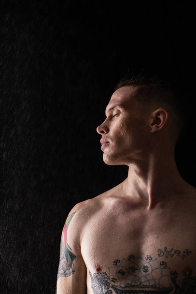 Tattooed man with water drops on his body, handsome healthy tattooed young man in the rain. Sexy fitness man. Naked torso with tattoos. Fashion male posing in studio on dark background. - Photo, image
