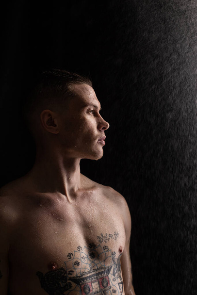 Homme tatoué avec des gouttes d'eau sur son corps, beau jeune homme tatoué en bonne santé sous la pluie. Homme de fitness sexy. Torse nu avec tatouages. Mode mâle posant en studio sur fond sombre. - Photo, image