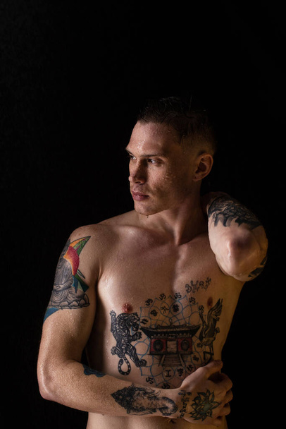 Tattooed man with water drops on his body, handsome healthy tattooed young man in the rain. Sexy fitness man. Naked torso with tattoos. Fashion male posing in studio on dark background. - Photo, Image