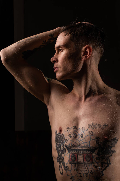 Hombre tatuado con gotas de agua en su cuerpo, guapo y saludable hombre joven tatuado bajo la lluvia. Hombre sexy de fitness. Torso desnudo con tatuajes. Moda masculina posando en estudio sobre fondo oscuro. - Foto, imagen