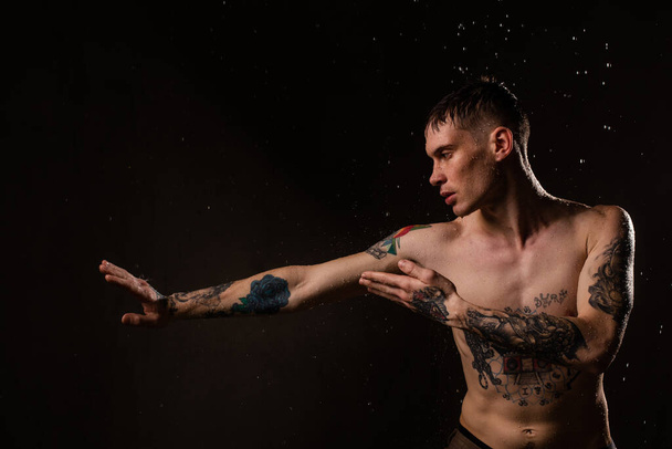 Tattooed man with water drops on his body, handsome healthy tattooed young man in the rain. Sexy fitness man. Naked torso with tattoos. Fashion male posing in studio on dark background. - Foto, afbeelding