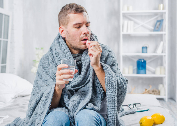 sick man wrapped scarf sitting bed taking pill with water. High resolution photo - Photo, Image