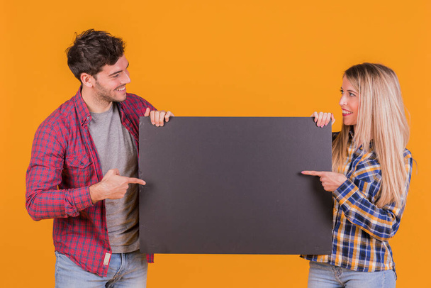 Porträt eines jungen Paares, das mit dem Finger auf ein schwarzes Plakat vor orangefarbenem Hintergrund zeigt. Foto in hoher Auflösung - Foto, Bild
