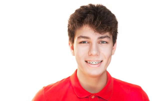 Nuori poika punaisessa paidassa valkoisella taustalla hymyilee hauskasti. Hän on iloinen, että hänellä on jopa kauniit hampaat, koska hänellä on hammasraudat hampaissaan. Eristetty. Kopioi tila - Valokuva, kuva