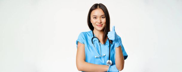 Enfermeira sorridente, médica asiática em esfoliação, mostrando os polegares para cima sinal e braço vacinado com gesso médico, recomendando a vacinação de covid-19, fundo branco. - Foto, Imagem
