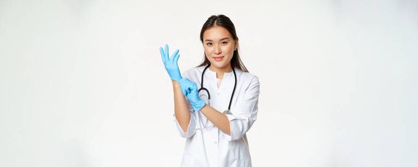 Lächelnd selbstbewusste asiatische Ärztin zieht medizinische Handschuhe an, bereitet sich auf die Untersuchung vor, steht in Uniform vor weißem Hintergrund. - Foto, Bild