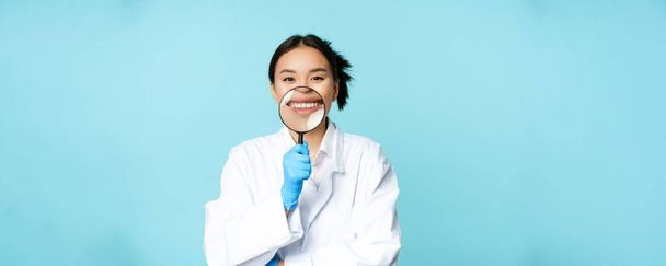 Funny asiática médica, médico mostrando su sonrisa con lupa, de pie en uniforme médico sobre fondo azul. - Foto, Imagen