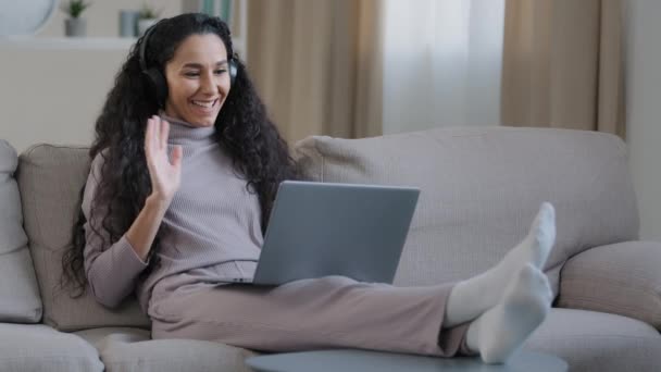 Hispánský arabský roztomilý žena nosit sluchátka sedět na pohovce doma obývací pokoj držet notebook použít video hovor vzdálenost vlna ruka pozdravit přítel usmívající se sociální sítě komunikace během pandemie - Záběry, video