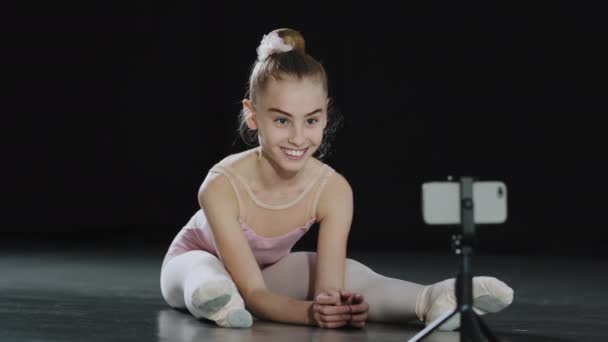 Genç kız çocuk balerin akrobat jimnastikçisi yerde oturup webcam 'e bakıyor. İnternette dans dersi veriyor. İnternet eğitmeni akıllı telefondan tek işaret parmağıyla işaret ediyor. - Video, Çekim