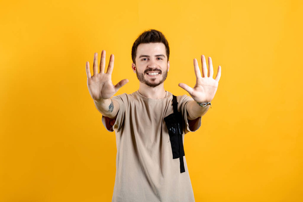 Jeune homme souriant confiant portant t-shirt beige posant isolé sur fond jaune montrant et pointant vers le haut avec les doigts numéro dix tout en souriant. - Photo, image