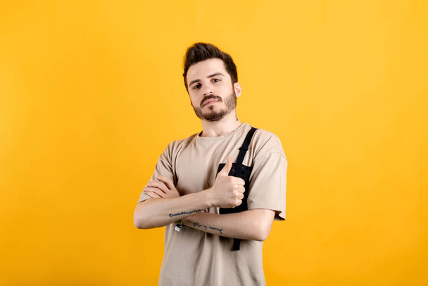 Handsome caucasian man wearing beige t-shirt posing isolated over yellow background arms crossed, looking at camera, friendly smiling, thumb up gesture - Photo, Image
