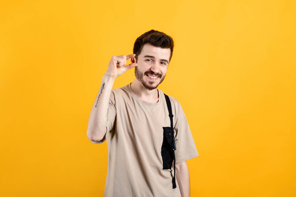 Joyeux jeune homme portant t-shirt posant isolé sur fond jaune en regardant la caméra montrant la mesure avec les doigts. Affichage de la petite longueur de taille avec une pitié. - Photo, image