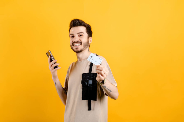 Wesoły biały mężczyzna ubrany w luźne ubrania, pozujący na żółtym tle z kartą kredytową lub debetową i smartfonem w rękach, wolna przestrzeń. - Zdjęcie, obraz
