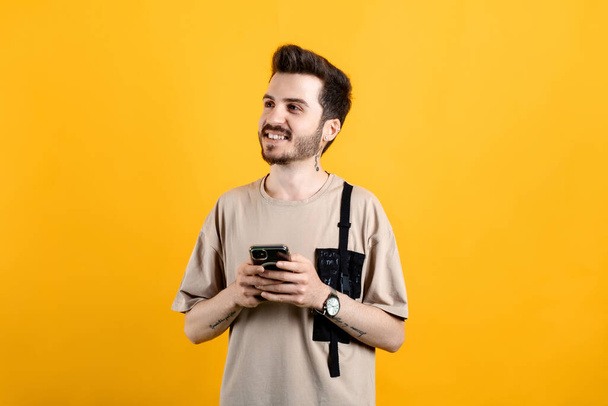 Cheerful young man wearing t-shirt posing isolated over yellow background dreamy look, thinking while holding smartphone and looking aside. - Photo, Image