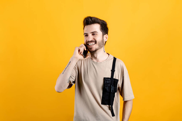 Nuori mies hymyilee itsevarma yllään beige tee poseeraa yli keltainen tausta puhuu älypuhelimella ja hymyilee. Hyvien uutisten vastaanottaminen puhelinkeskustelun aikana. - Valokuva, kuva
