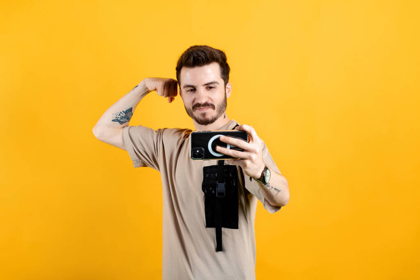 Komea nuori mies yllään rento vaatteita poseeraa yli keltainen tausta ottaen selfie, osoittaa vahva käsi, taipuisa hauis ja poseeraa valokuva. - Valokuva, kuva