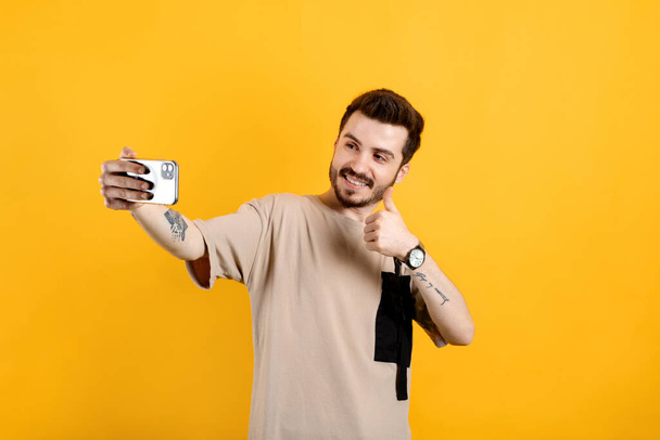 Beau jeune homme portant des vêtements décontractés posant isolé sur fond jaune faire un geste correct tout en prenant selfie ou avoir un appel vidéo. - Photo, image