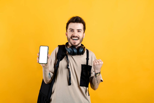 Sarı arka planda izole edilmiş bej tişört giyen mutlu genç adam kameraya beyaz ekranlı telefonu gösteriyor. Gülümseyen ve başarı için çığlık atan. - Fotoğraf, Görsel
