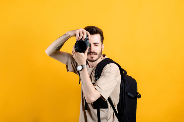 Valkoihoinen nuori mies t-paidassa poseeraa eristettynä keltaisen taustan päällä ottamassa kuvia dslr-kameralla. Valokuvaaja peittää kasvonsa kameralla. - Valokuva, kuva