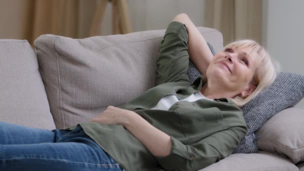 Onnellinen rento keski-ikäinen pensive haaveilee nainen levätä sohvalla kotona nauttia rauhallisella tuulella, uninen laiska rauhallinen vanha vanhempi kypsä nainen unelmia makaa mukava sohva olohuoneessa stressiä ilmaiseksi rentoutua - Materiaali, video