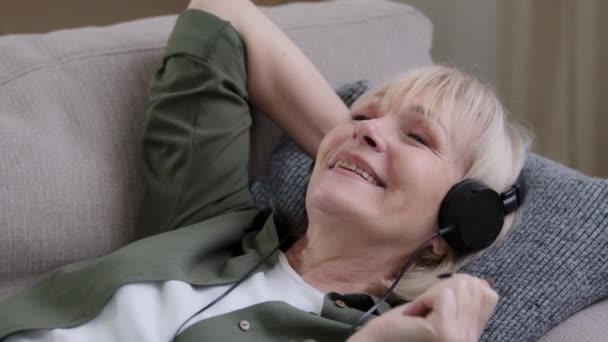 Zbliżenie starsza kobieta w słuchawkach słuchając muzyki leżącej na soga w domu. zrelaksowany stary kaukaski lady w słuchawki słuchać dźwięk piosenka szczęśliwy beztroski babcia spoczywa na kanapie śpiew mając zabawy - Materiał filmowy, wideo