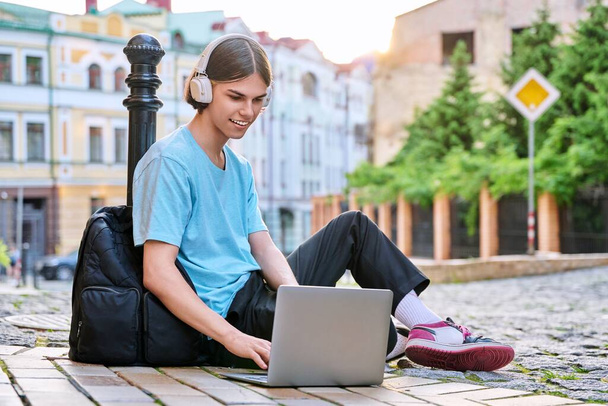 Teenage male student in headphones using laptop outdoor, on city street. Guy 17, 18 years old looking at screen, sitting on sidewalk. College, high school, youth, education, technology concept - Foto, Bild