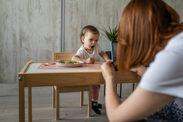 nieznana kobieta matka fotografowanie dziecko dziewczynka mały biały maluch jedzenie podczas siedzieć przy stole w domu kopia przestrzeń małe dziecko jedzenie obiad zdrowy posiłek w pokoju w dzień poważne skoncentrowane - Zdjęcie, obraz