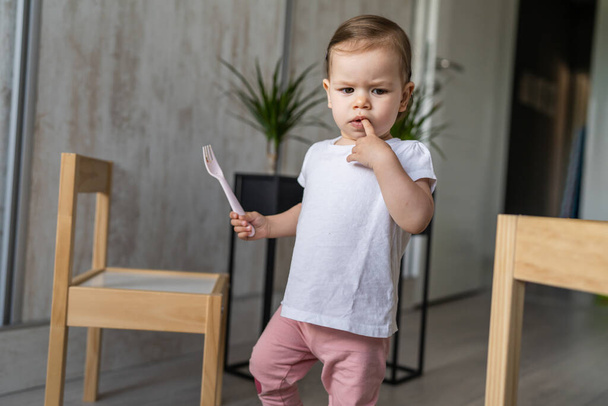 una niña pequeña niña caucásica pequeño niño caminando en la habitación en casa en el día mantenga tenedor de plástico con el dedo en su boca copia espacio serio reflexivo - Foto, imagen