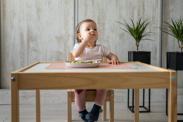 One baby girl small caucasian toddler eating while sit at the table alone at home copy space little child having lunch healthy food meal in room in day front view - Φωτογραφία, εικόνα