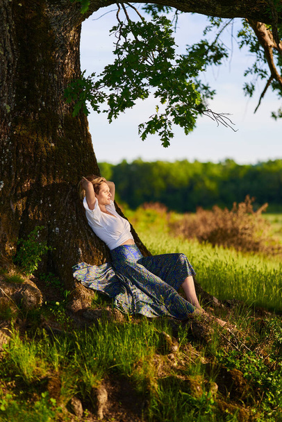 Attractive young blonde woman posing by a huge centennial oak tree at sunset - Foto, Imagem