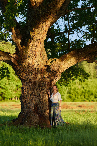 Attractive young blonde woman posing by a huge centennial oak tree at sunset - Fotó, kép