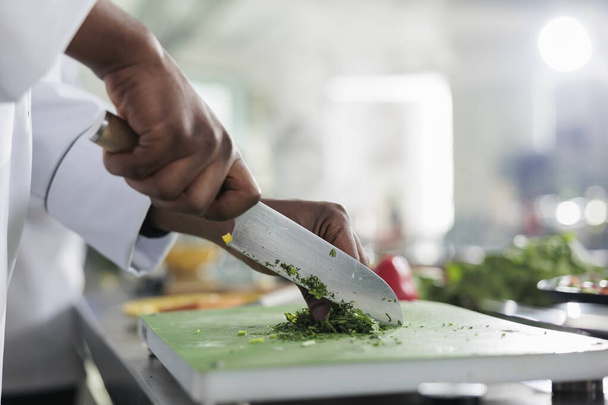 Primer plano de las manos afroamericanas utilizando cuchillo profesional para cortar hierbas y verduras orgánicas finamente frescas para la receta de guiso. Cocinar corte de verduras para el plato gourmet servido en el restaurante de alta cocina. - Foto, imagen